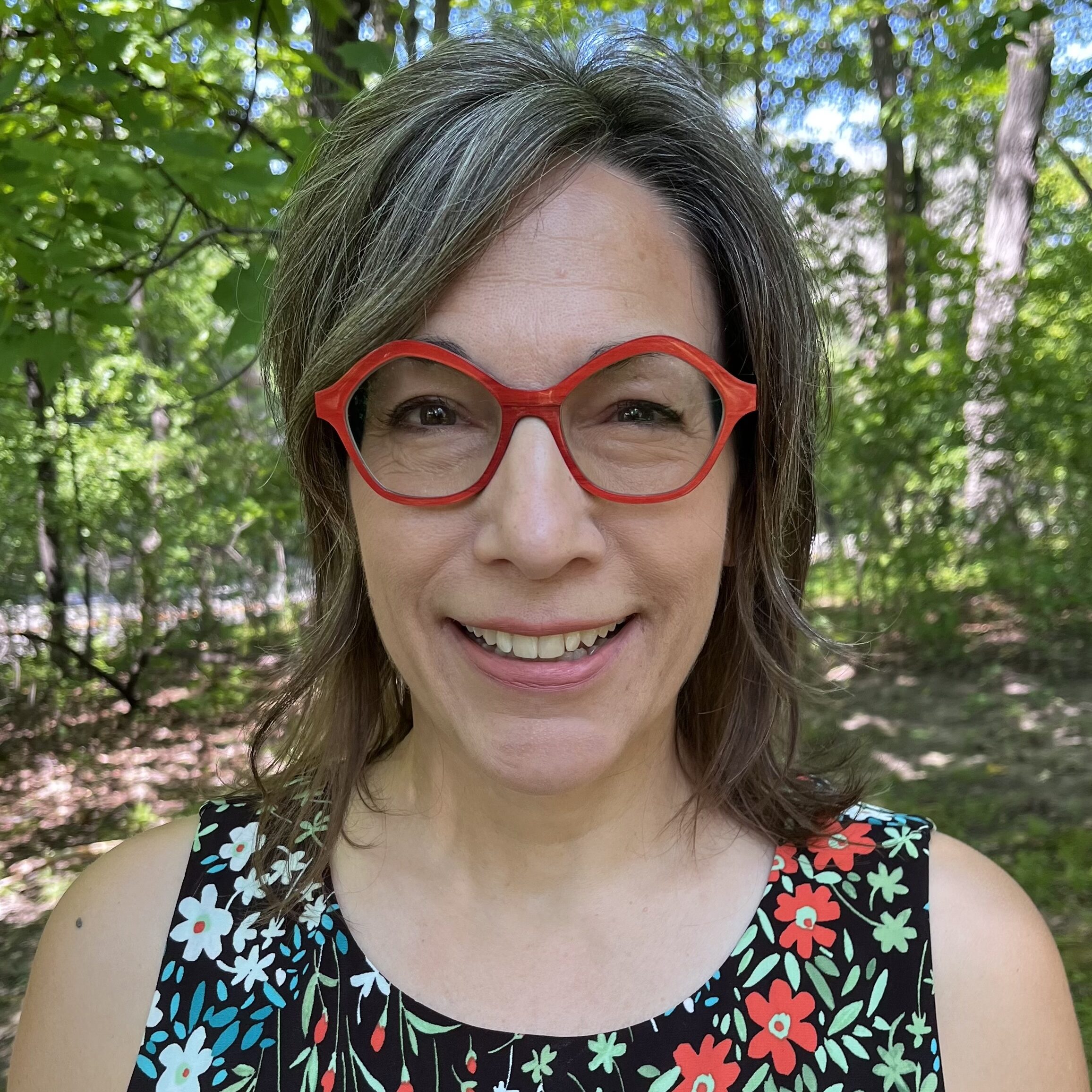 Portrait of Heidi Marty wearing red glasses and a floral top standing outside with trees in the background.